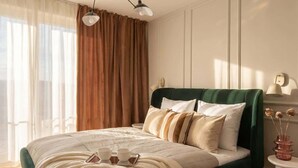 Une chambre moderne avec un lit recouvert d’un tissu vert, de nombreux coussins, une table de chevet avec un vase et une grande fenêtre avec des rideaux.