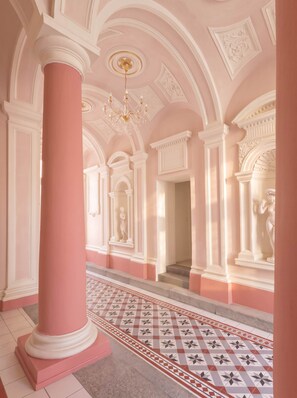 A grand hallway with a pink and white color scheme, featuring a large column, a patterned floor, and classical architectural details.