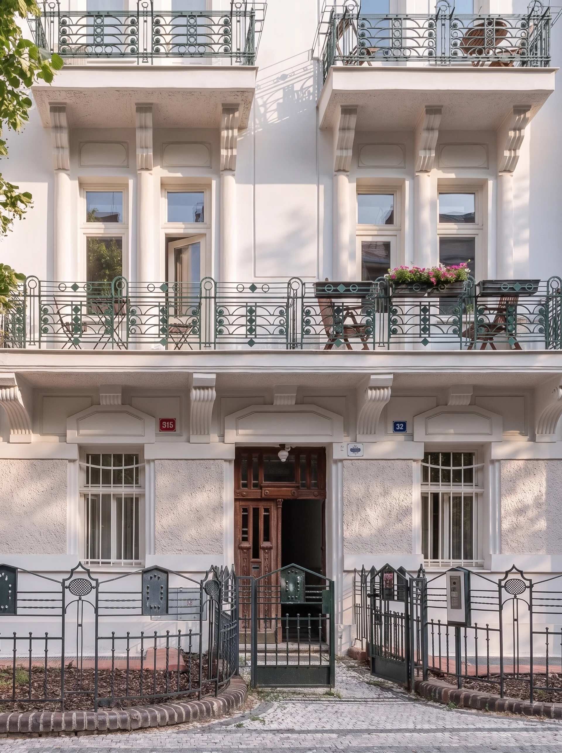 Ein mehrstöckiges Gebäude im klassischen Stil, mit einem zentralen Eingang mit Holztür, einem Balkon mit Metallgeländern und einer kleinen Gartenanlage mit einer Bank.