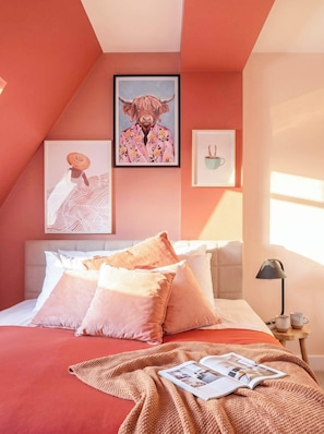 A bedroom with a sloped ceiling, a bed with pink bedding, and artwork on the walls.