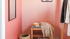 A room with a pink wall, a mirror, two framed artworks, a wooden shelf with books and a blanket, a woven basket, and a pair of shoes.