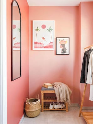A room with a pink wall, a mirror, two framed artworks, a wooden shelf with books and a blanket, a woven basket, and a pair of shoes.