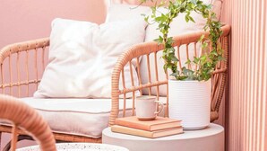 A cozy corner with a wicker chair, a white pillow, a small potted plant, a stack of books, and a white mug on a round table.