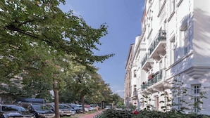 A row of white multi-story buildings with balconies, parked cars, and trees along the street.