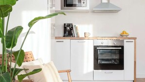 Eine moderne Küche mit weißen Schränken, einer integrierten Mikrowelle, einer Dunstabzugshaube und einem Backofen. Ein Holzstuhl mit Kissen, ein Topfpflanze und ein Teppich liegen auf dem Boden.