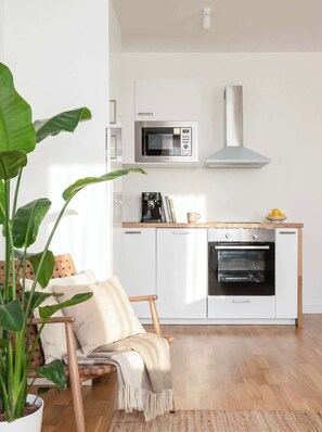 A modern kitchen with white cabinetry, a built-in microwave, a range hood, and an oven. There is a wooden chair with a cushion, a potted plant, and a woven rug on the floor. - TurnKey | National Museum Apartment V (Praha 1)