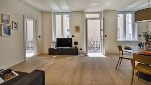 Ein modernes Wohnzimmer mit Flachbildfernseher, einer Couch, einem Esstisch mit Stühlen und großen Fenstern mit Blick nach außen.