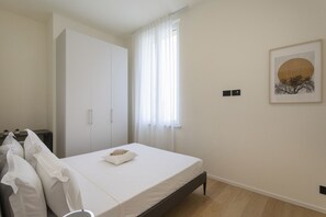 Een slaapkamer met een bed, een kledingkast, een raam met gordijnen, een nachtkastje en een lijst met een afbeelding aan de muur.