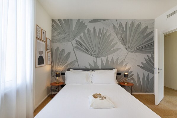 Een slaapkamer met een groot bed, twee nachtkastjes met lampen, een muur met een bladmotief en een deur naar een andere kamer.