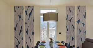 A dining area with a round table set for six, surrounded by black chairs. The walls feature a leaf pattern wallpaper, and a large window offers a view of buildings outside. - Riviera Flavour Apartments by Wonderful Italy - Timo (Santa Margherita Ligure)