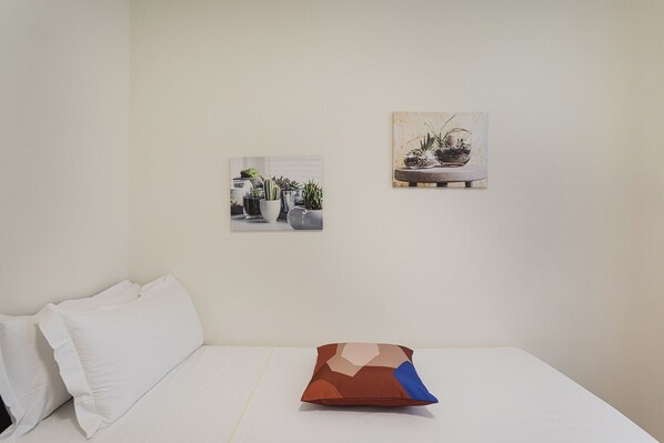 Un dormitorio con una cama blanca, un almohadón y dos cuadros en la pared.