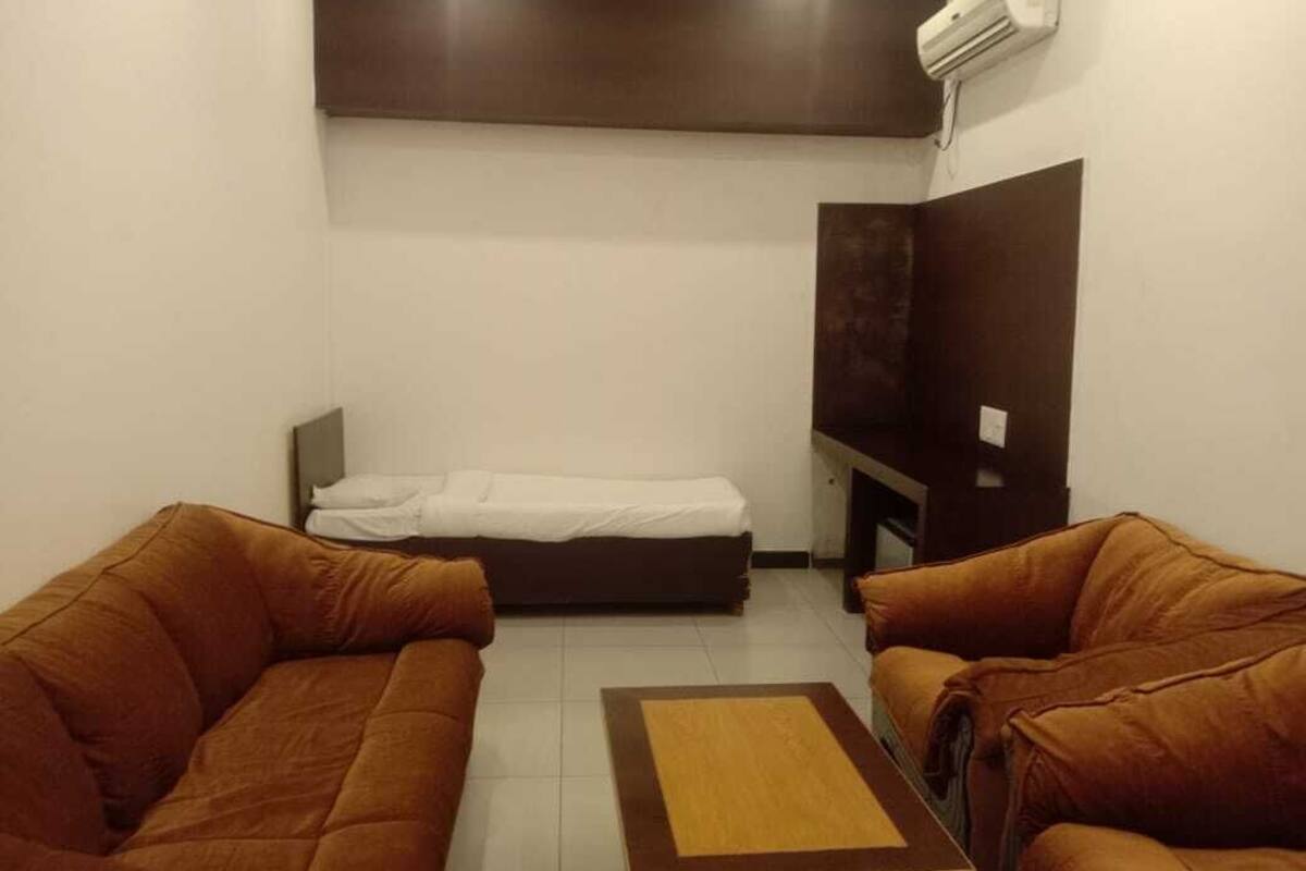 A small, furnished room with two brown sofas, a single bed, a flat-screen TV, and a wall-mounted air conditioner.