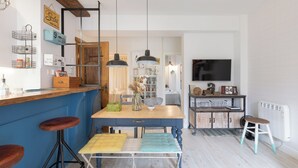 A modern kitchen with a blue bar counter, wooden stools, a dining table with a bench, pendant lights, and a television mounted on the wall. - Astoria - baskeyrentals (San Sebastián)
