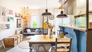A cozy living room with a sofa, a wooden dining table, and a kitchen area with a fridge and shelves. - Astoria - baskeyrentals (San Sebastián)