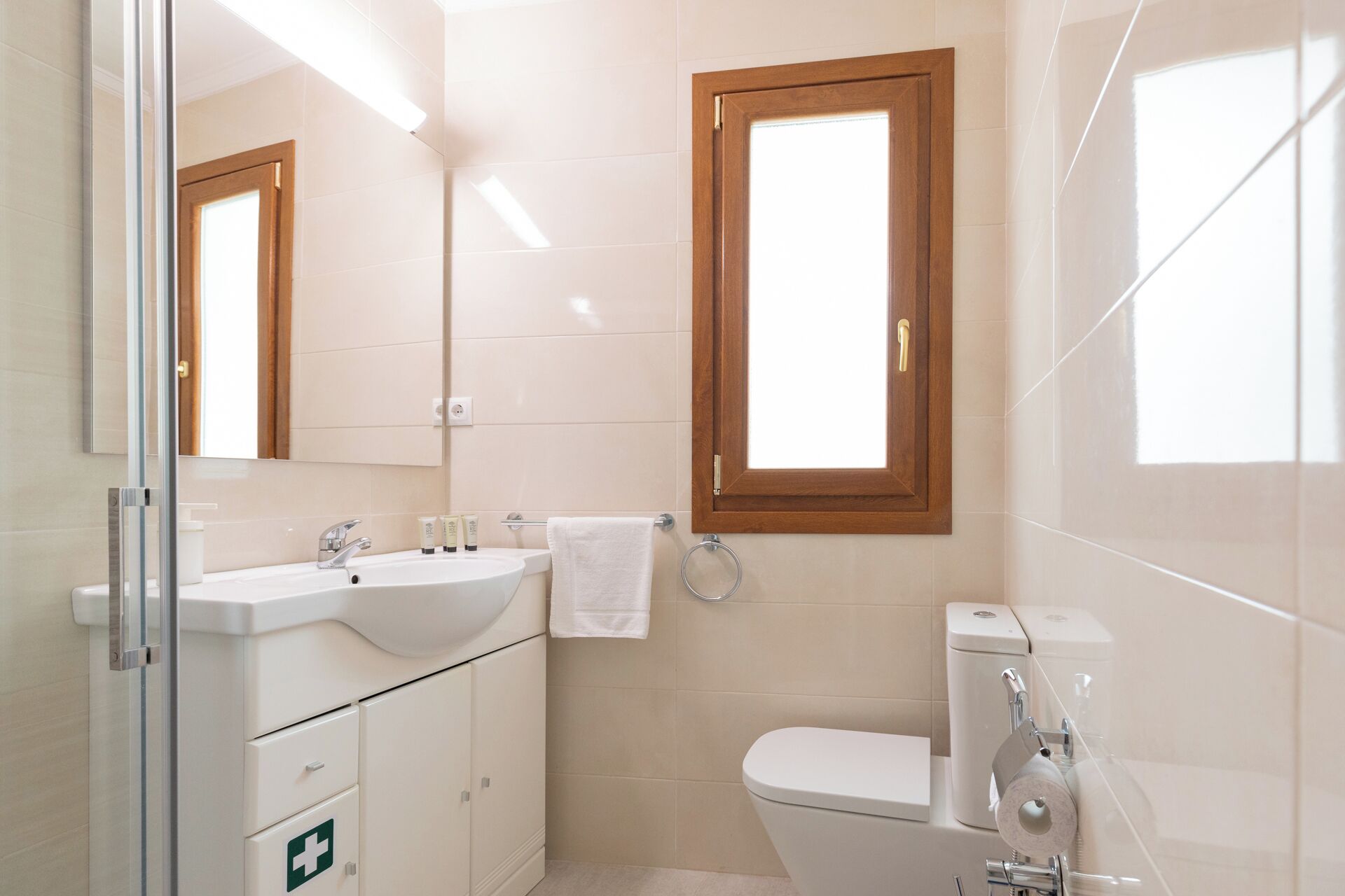 Une salle de bain moderne avec un lavabo blanc, un miroir et des toilettes. Une fenêtre à cadre en bois se trouve au-dessus des toilettes, et la douche est dotée d’une porte en verre.