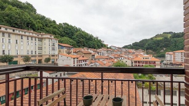 A balcony with wooden railings overlooking a town with terracotta-roofed buildings and lush green hills.