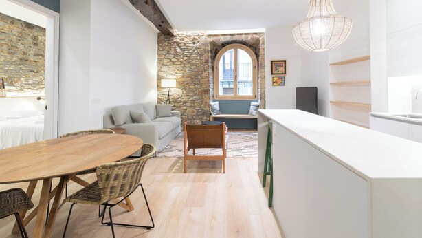A modern kitchen with a stone wall, a dining area with a wooden table and wicker chairs, and a view of the city through a window. - Zutabe - baskeyrentals (Azpeitia)