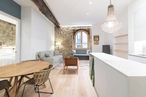 A modern kitchen with a stone wall, a dining area with a wooden table and wicker chairs, and a view of the city through a window. - Zutabe - baskeyrentals (Azpeitia)