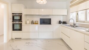 A modern kitchen with white cabinetry, a built-in oven, a microwave, a kettle, and a large window providing natural light.