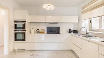 A modern kitchen with white cabinetry, a built-in oven, a microwave, a kettle, and a large window providing natural light.