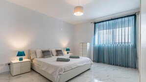 A modern bedroom with a large bed, two bedside tables with lamps, a nightstand, and a window with blue curtains.