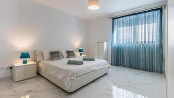 A modern bedroom with a large bed, two bedside tables with lamps, a nightstand, and a window with blue curtains.