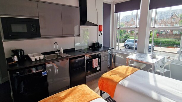 A hotel room with a kitchenette, two beds with orange blankets, a dining table, and a view of houses through large windows.