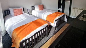 A bedroom with two beds, each with an orange blanket, white bedding, and patterned pillows. There is a small bedside table with a lamp, a radiator, and a framed picture on the wall.