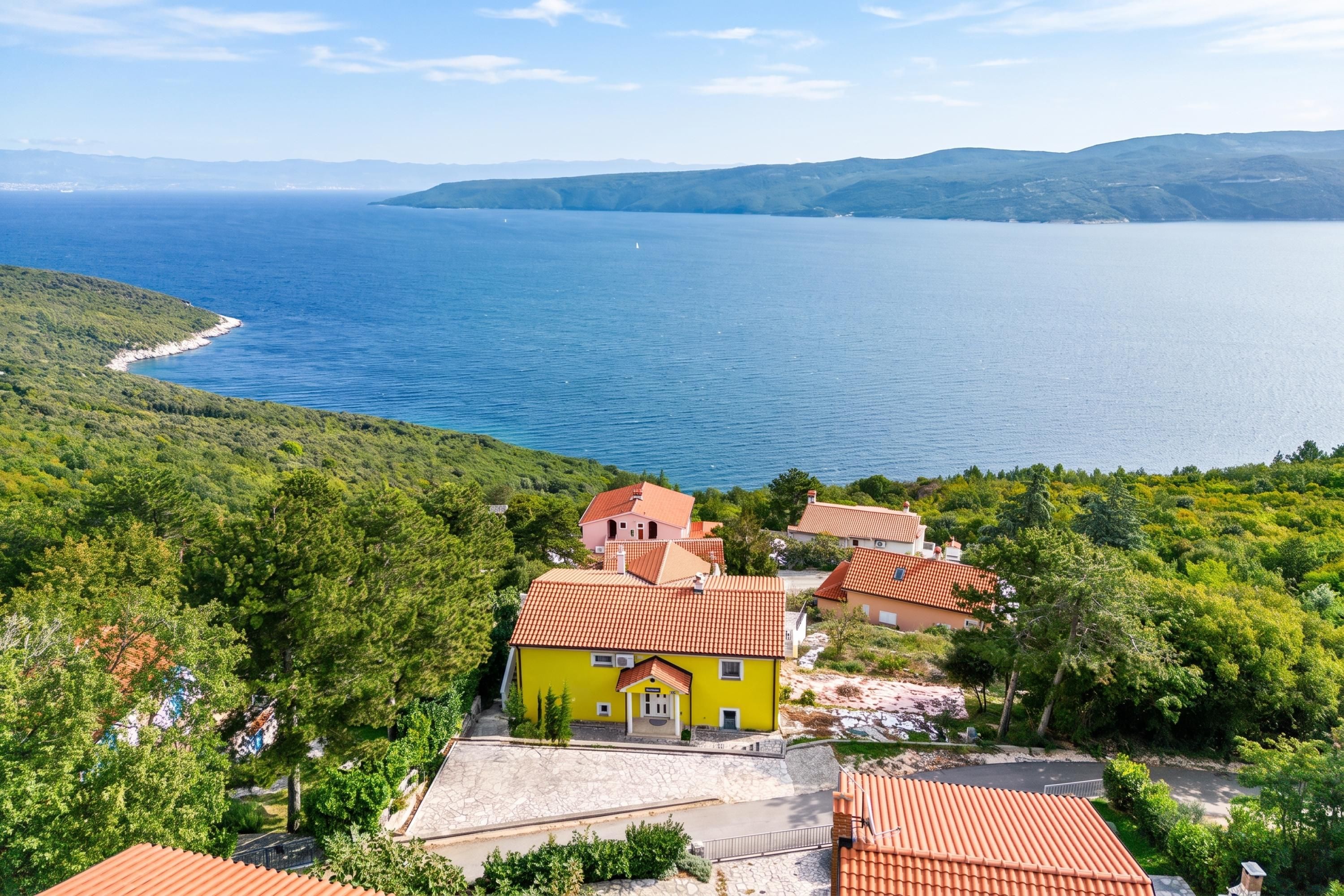 A coastal property with a yellow building, red-tiled roofs, and a view of the sea and hills.