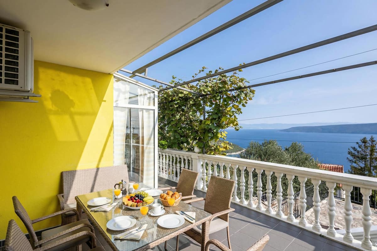 Un balcón con mesa lista para el desayuno, vista al mar y una pared amarilla.