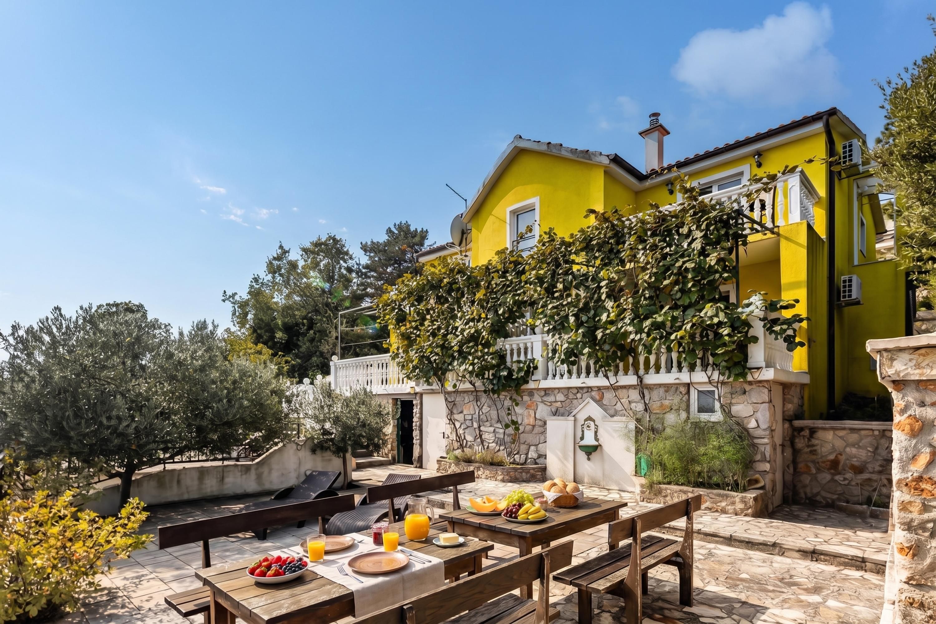 Une villa jaune à la façade en pierre, dotée d’un espace repas extérieur avec des tables et des bancs en bois, et offrant une vue sur la verdure environnante.