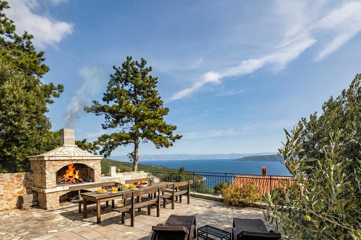 Un patio exterior con horno de pizza de piedra, mobiliario de madera y vista al mar.