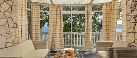 A sunroom with wicker furniture, a glass table with a tray of pastries, and a view of greenery outside.