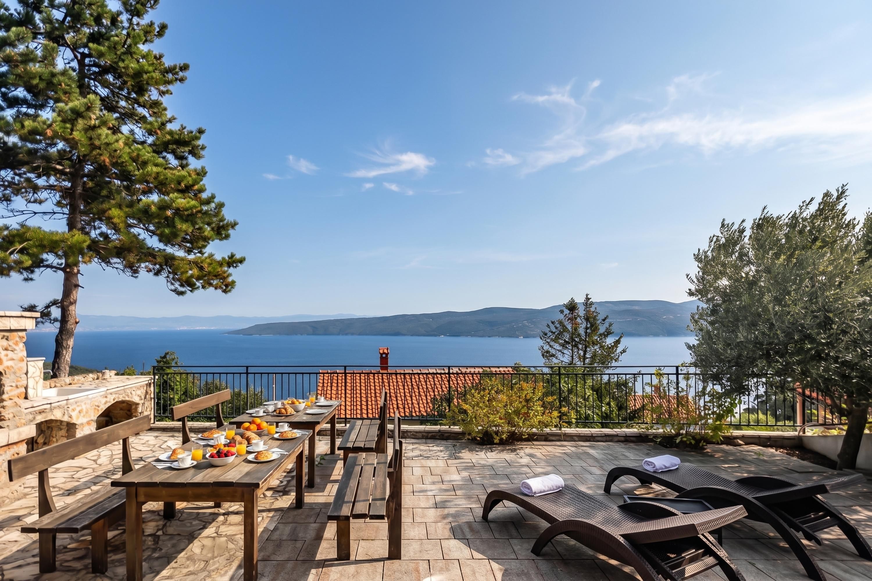 An outdoor dining area with stone flooring, wooden tables, and chairs, overlooking a body of water and distant hills.