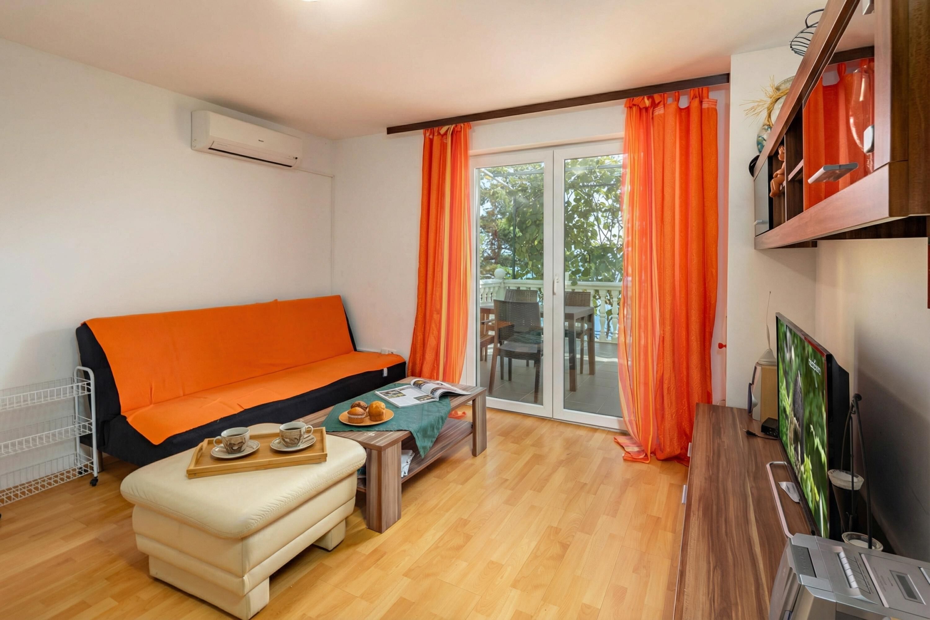 Sala de estar com sofá, mesa de centro com bandeja de doces, televisão e porta de vidro deslizante com cortinas.