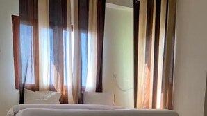 A bed with a wooden frame and white bedding in a room with a tiled floor and sheer curtains. - Earth View Residency  (Noida)