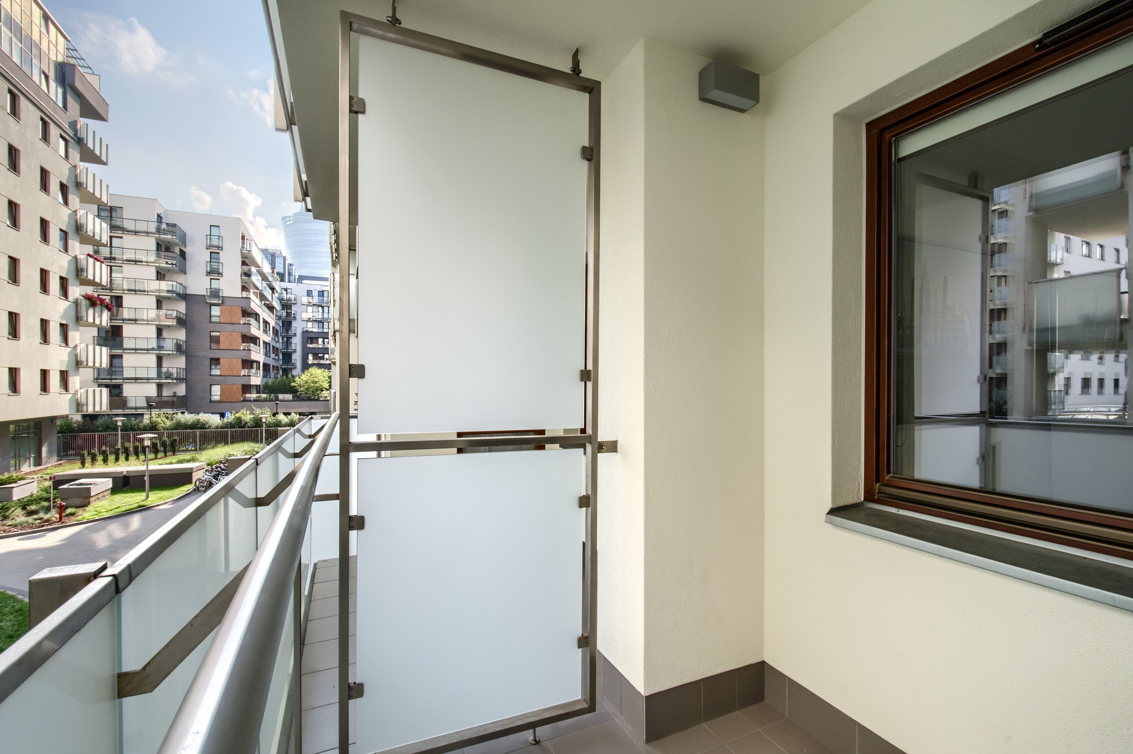 A balcony with a metal railing, a window with a wooden frame, and a view of a cityscape with modern buildings.