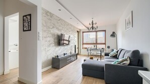 A modern living room with a grey sofa, a TV mounted on the wall, and a dining area with a table and chairs. - Warsaw Night Apartments Giełdowa (Warsaw)