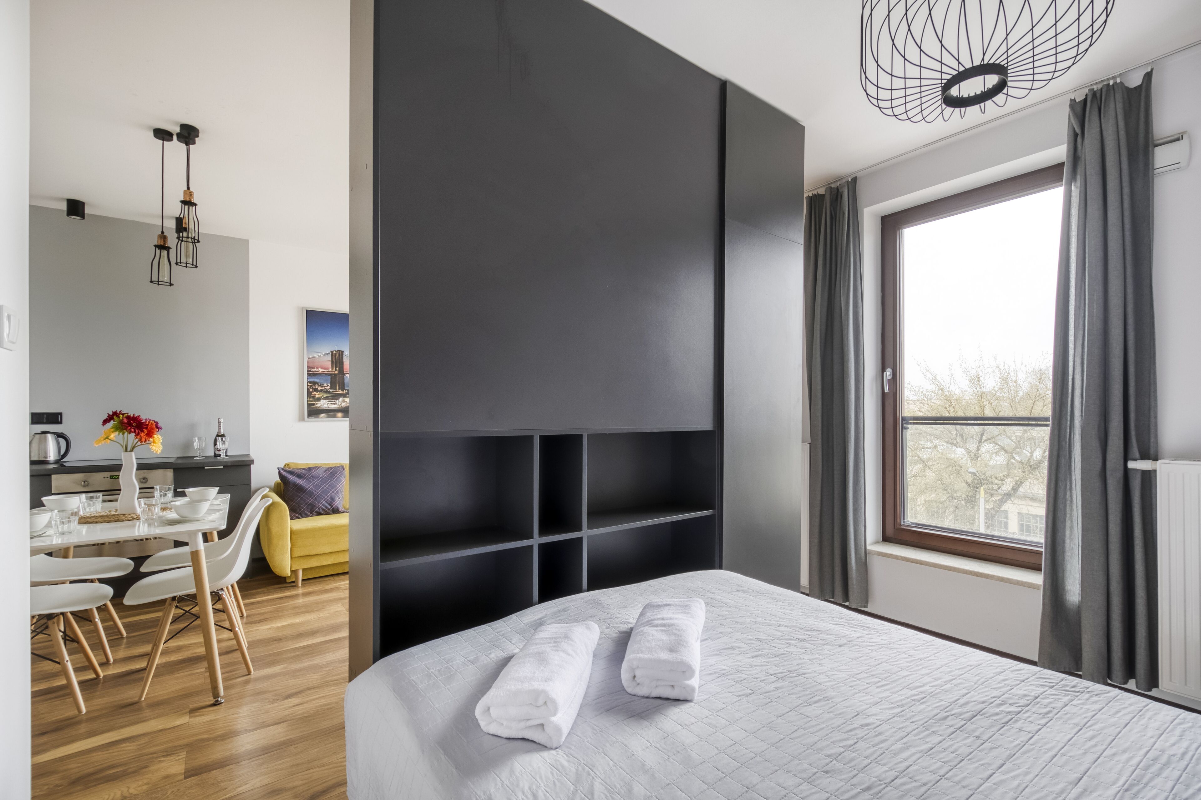 Une chambre d’hôtel moderne avec un grand lit, un coin repas comprenant une table et des chaises blanches, et une vue sur la ville par la fenêtre.