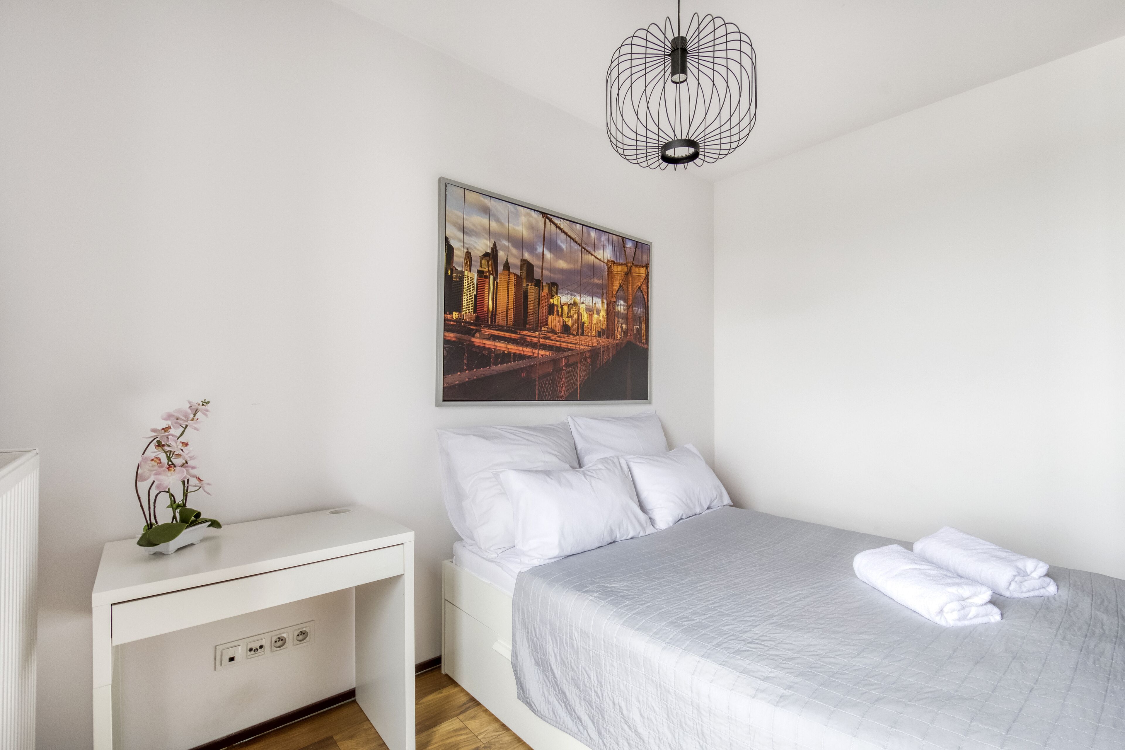 Une chambre à coucher avec un lit, une table de chevet blanche, un tableau représentant un paysage urbain accroché au mur, et un luminaire suspendu moderne.
