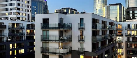 A multi-story residential building with balconies, a parking area, and a view of the city skyline.