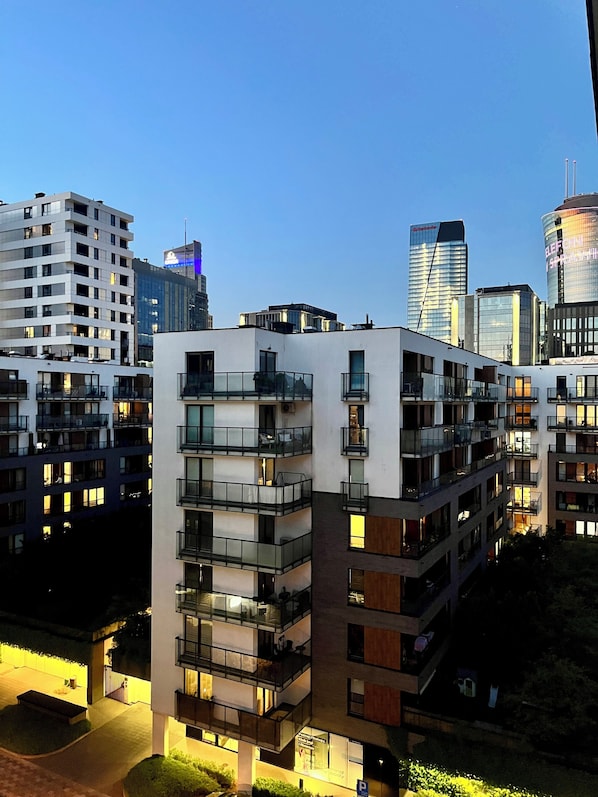 A multi-story residential building with balconies, a parking area, and a view of the city skyline.