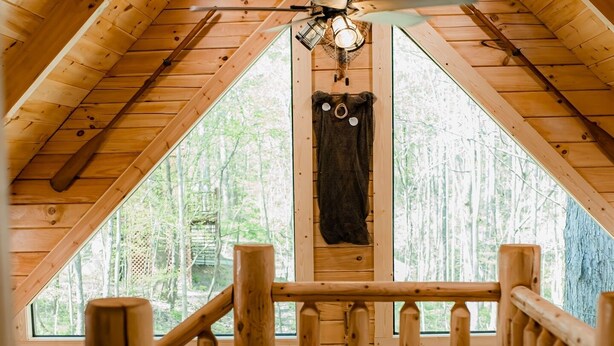 Un intérieur en bois avec un plafond en pente, un ventilateur de plafond central et une vue sur la forêt par une grande fenêtre.