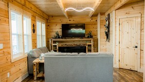 L'intérieur d'un chalet confortable, avec un canapé gris, des murs en bois, une cheminée et un téléviseur fixé au-dessus.