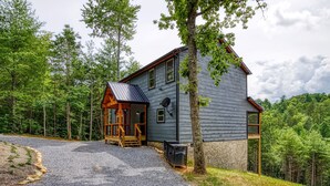A two-story cabin with a wooden porch and a gravel driveway, surrounded by a forested area.