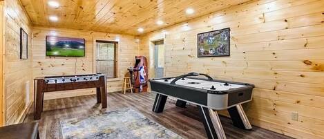Una habitación con piso de madera, dos mesas de fútbol de mesa, un banco y un televisor montado en la pared.