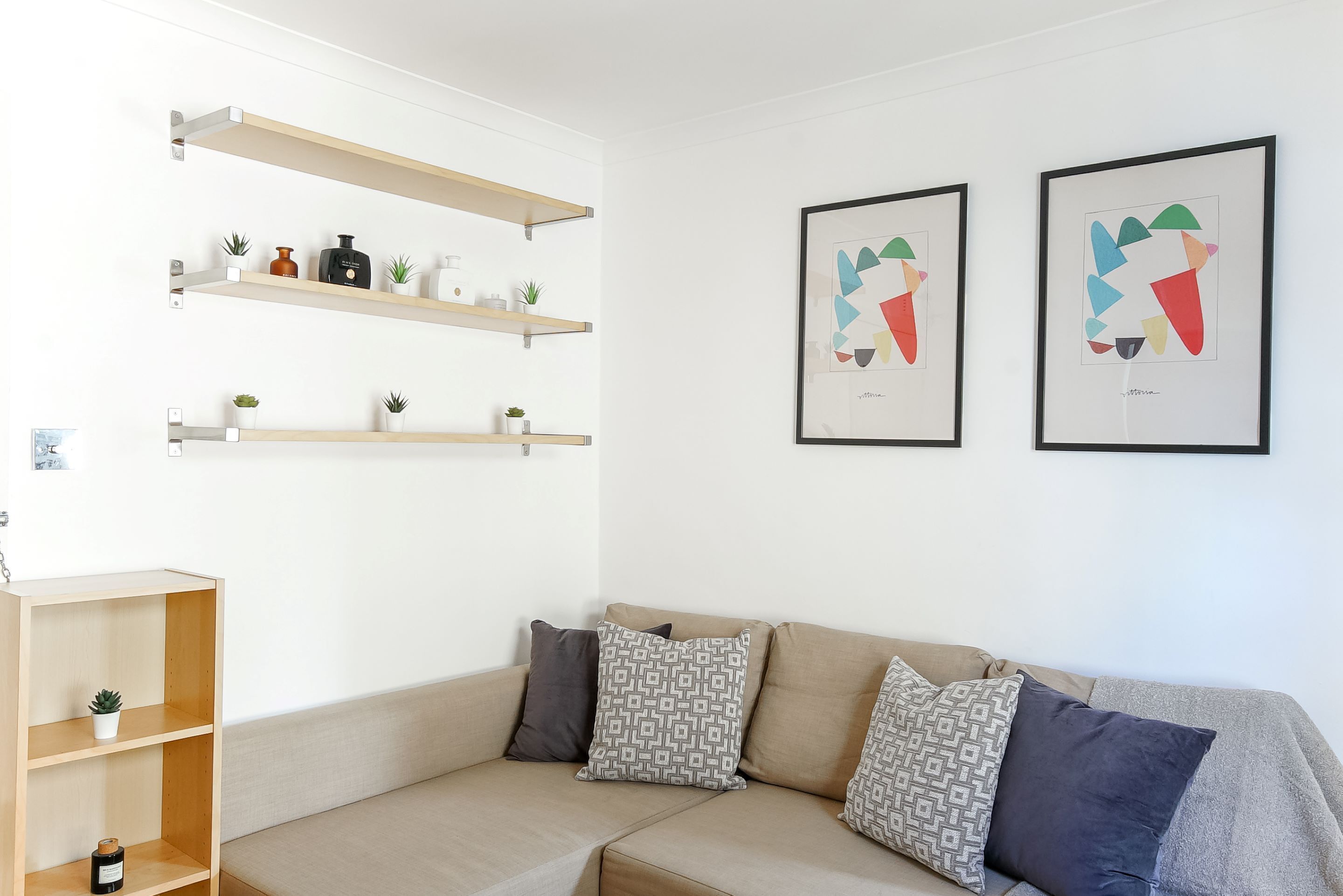 A modern living room with a beige sectional sofa, two framed abstract paintings, and wooden shelves with decorative items.