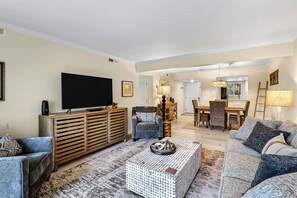 Una sala de estar con televisor de pantalla plana, mueble de madera para entretenimiento, dos sofás, una mesa de centro y un comedor con mesa y sillas.