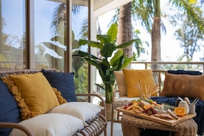 A wicker sofa with cushions, a wooden tray with fruits and snacks, and a view of palm trees through large windows. - Kalani On Airlie (Airlie Beach)
