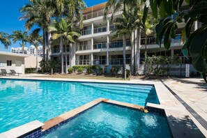 A swimming pool with a clear blue water, surrounded by a white building with multiple balconies, palm trees, and a fenced area. - Kalani On Airlie (Airlie Beach)